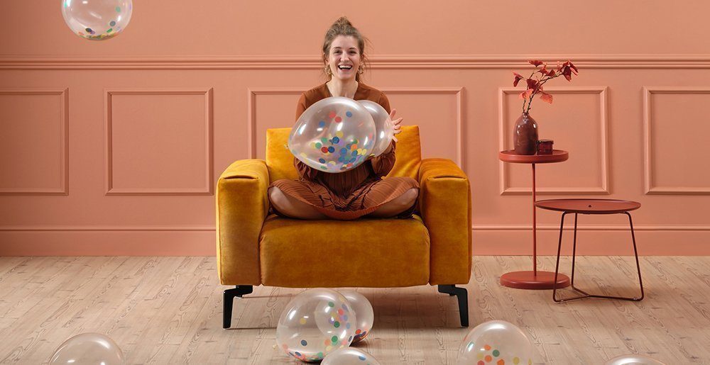 Sensoo – einfach den Komfort einstellen Eine junge Frau sitzt im Schneidersitz auf einem gelben Cosy1 1-Sitzer und spielt mit Luftballons. Dabei lacht sie. Das soll zeigen wie spielend einfach der Komfort bei Sensoo angepasst werden kann.